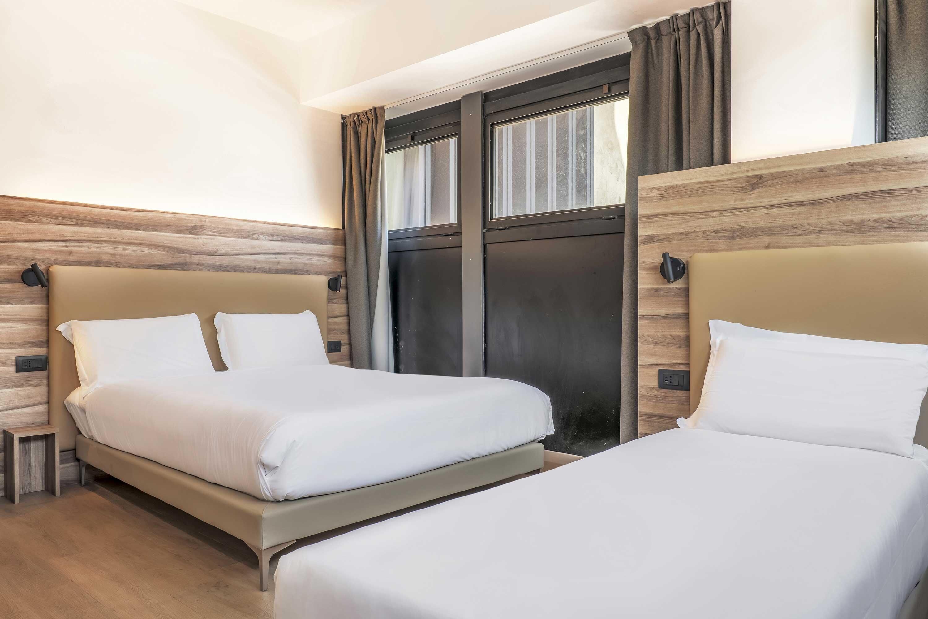 A hotel room with two beds, wooden paneling on the walls, a window with curtains, and a small bedside table.