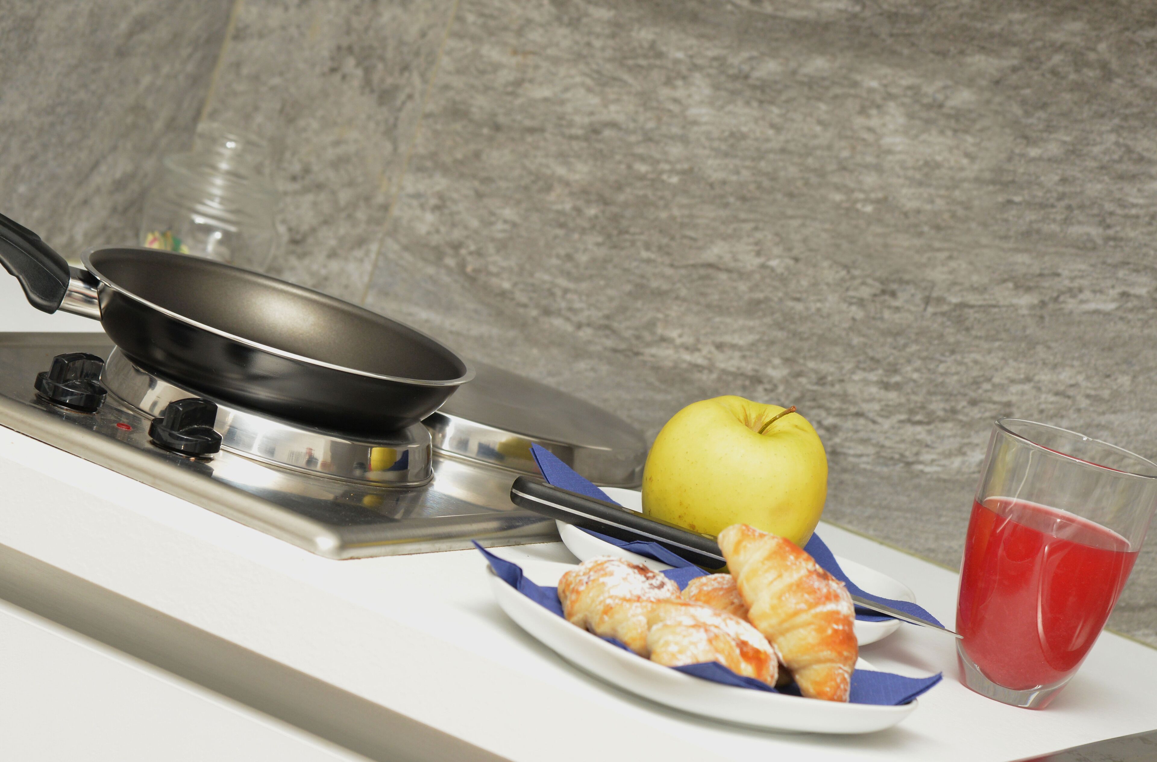 Un plan de travail de cuisine avec une poêle, une pomme, un verre de jus rouge et une assiette de pâtisseries.