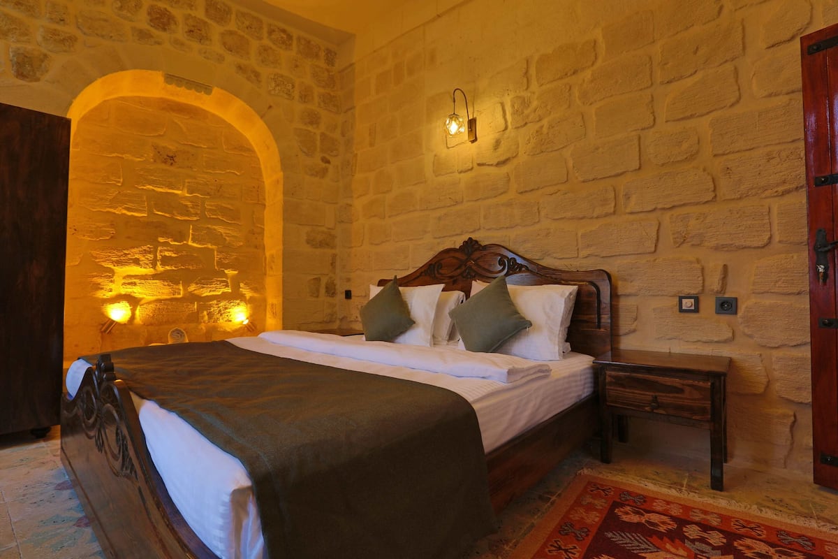 A bedroom with a stone wall, a wooden bed with a headboard, a nightstand, and a red patterned rug.