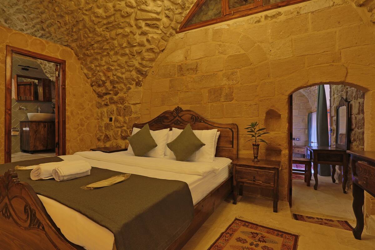 A stone-walled bedroom with a wooden bed, a nightstand, a mirror, and a view of the bathroom.