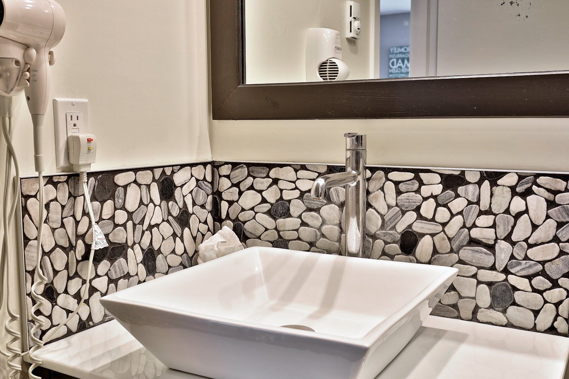 A bathroom with a white sink, a stone mosaic backsplash, a modern faucet, and a hairdryer plugged in.