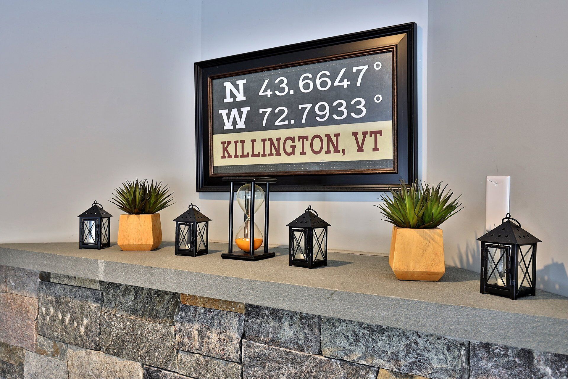 A framed sign with coordinates and location, flanked by potted plants and lanterns on a stone ledge.