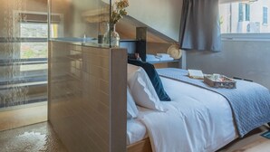 Un dormitorio moderno con cabina de ducha de vidrio, cama con ropa de cama blanca, mesita de noche con un florero y vistas a la calle a través de una ventana.
