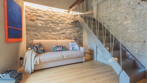 Una sala acogedora con pared de piedra, un sofá color beige, cojines de colores, una máquina de escribir antigua y una escalera con barandilla de vidrio.