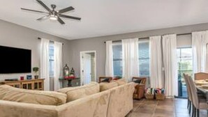 A living room with a beige sectional sofa, a ceiling fan, a flat-screen TV, and a dining area with a table and chairs. - CG 9025 Shadow Mountain Epic Villa By Disney Theater Game Room Pool Spa (Davenport)