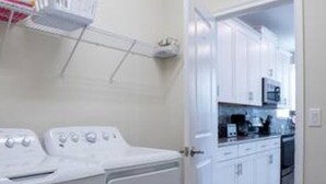 A laundry area with a washing machine and dryer, a basket of folded clothes, and a kitchen visible through an open door. - CG 1557 Mulligan Magical Retreat 5BR Pool Spa Near Disney (Davenport)