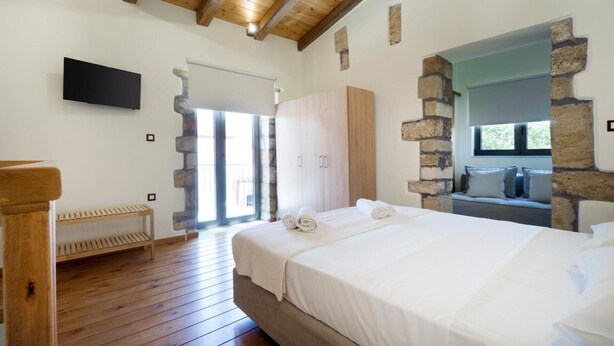 A bedroom with a wooden ceiling, stone walls, a bed with white linens, a bench, and a TV on the wall. - Kyriaki's Traditional House (Chania)