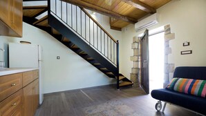 A kitchen with wooden cabinets, a refrigerator, and a patterned rug. A staircase with a black railing leads to a stone wall. - Kyriaki's Traditional House (Chania)