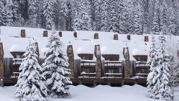 Een rij houten blokhutten met sneeuw bedekte daken en bomen ervoor.