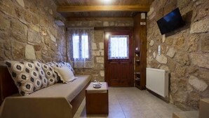 A cozy room with a stone wall, a wooden ceiling, a sofa with patterned cushions, a small table with a tray, a door with a window, and a mounted TV.