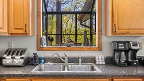 A kitchen with wooden cabinets, a double sink, a toaster, a coffee maker, and a window with a view of trees. - Pet-Friendly · 3 Kings · Hot Tub · Arcade + Pool Table (Gatlinburg)