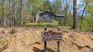 A cabin with a stone chimney, surrounded by trees and a dirt path leading to it. - Pet-Friendly · 3 Kings · Hot Tub · Arcade + Pool Table (Gatlinburg)