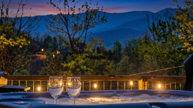 A hot tub with two glasses of water, surrounded by a wooden deck and a mountainous backdrop. - Pet-Friendly · 3 Kings · Hot Tub · Arcade + Pool Table (Gatlinburg)