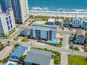 A multi-story hotel with a swimming pool, located near a beach with visible waves.