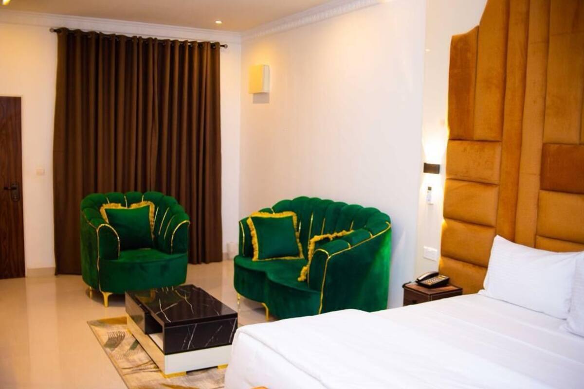 Habitación de hotel con una cama grande, dos sillones verdes, una mesita y un cabecero marrón.