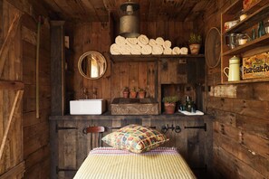 A rustic wooden room with a bed, a wooden cabinet with a mirror, a sink, and a shelf with various items.