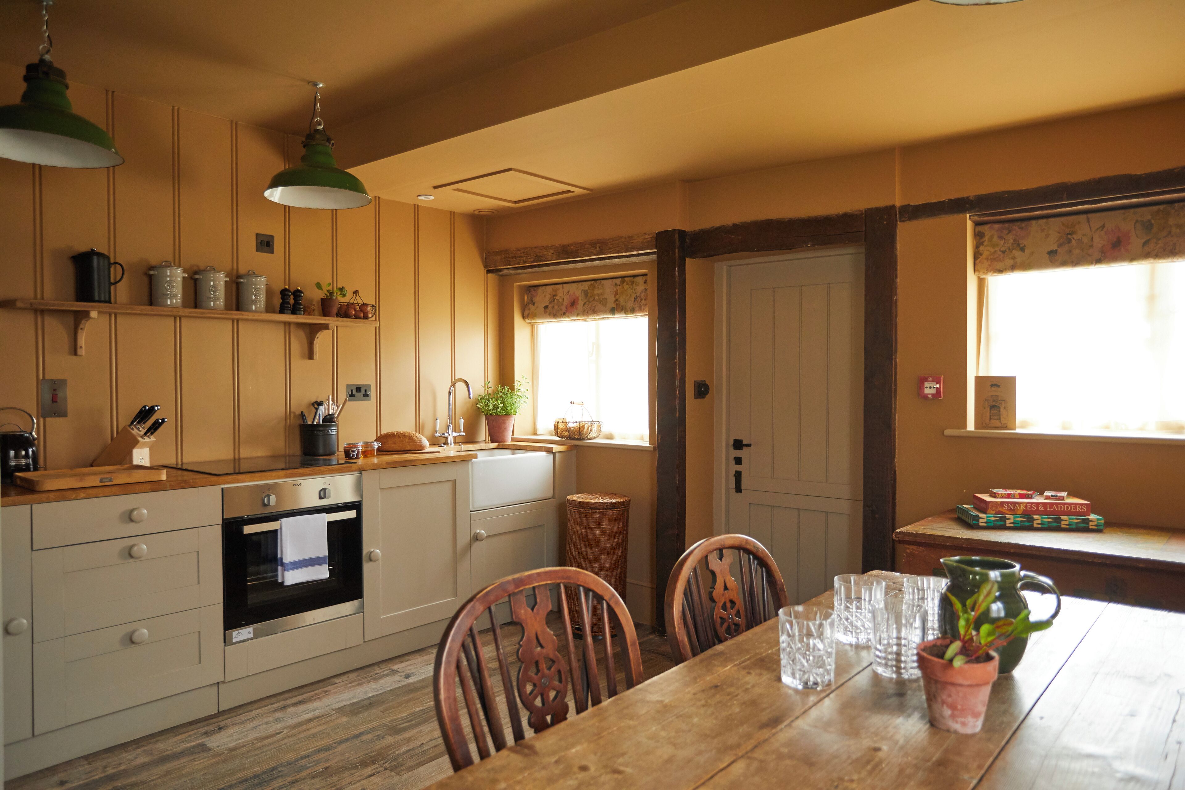 Una cocina con armarios de madera, una mesa de comedor con sillas, una ventana con cortinas florales y una estantería con utensilios de cocina.