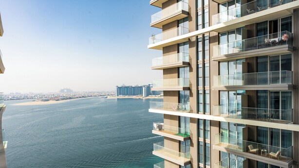 A high-rise building with multiple balconies overlooking a body of water and distant cityscape.