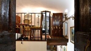 Un escalier avec une rampe en bois, une chaise et une table dans une pièce dont le sol est recouvert de carreaux et qui possède une porte ornée de motifs décoratifs.