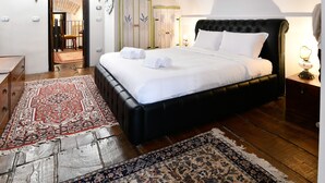 Une chambre avec un grand lit, une commode en bois, un tapis à motifs rouges et blancs, et un mur blanc orné d’un miroir décoratif.