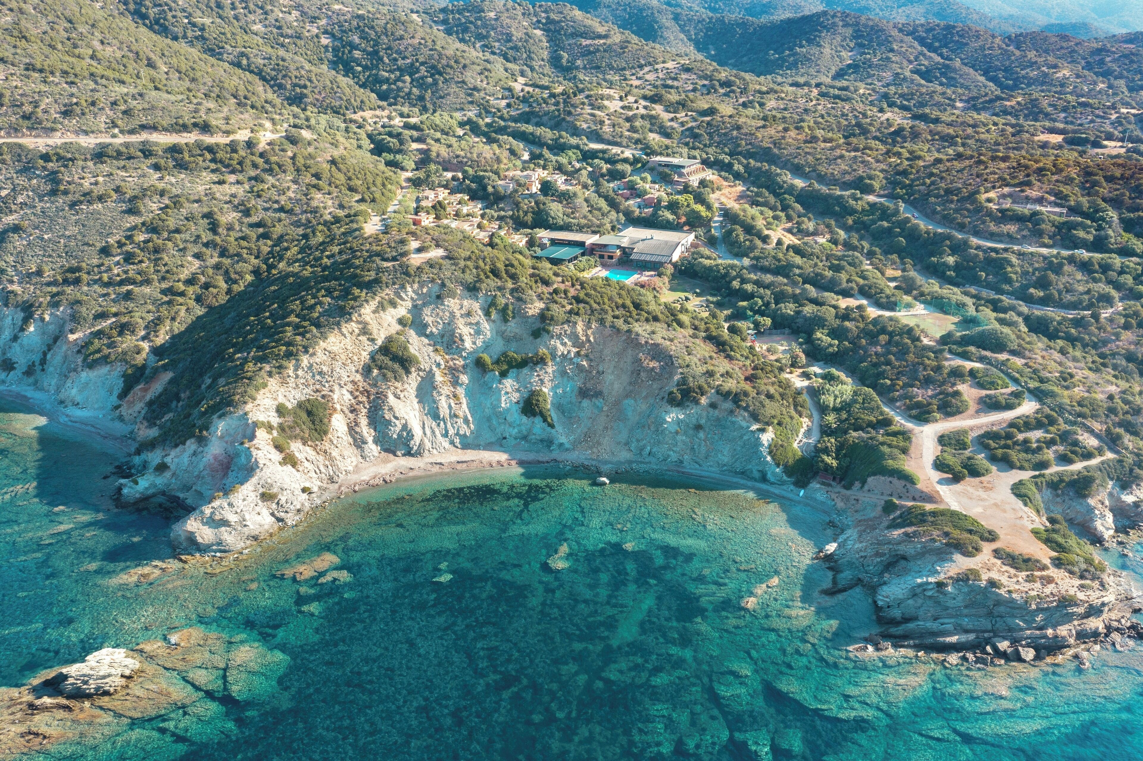An aerial view of a coastal property with a swimming pool, surrounded by rocky terrain and dense vegetation.