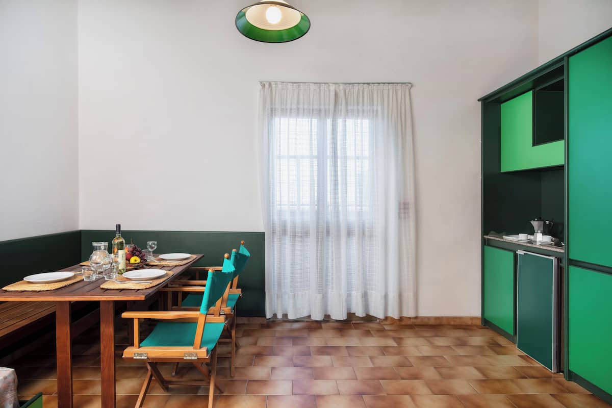 Un coin repas avec une table en bois dressée pour quatre personnes, des chaises, une fenêtre ornée de rideaux blancs et un buffet vert.
