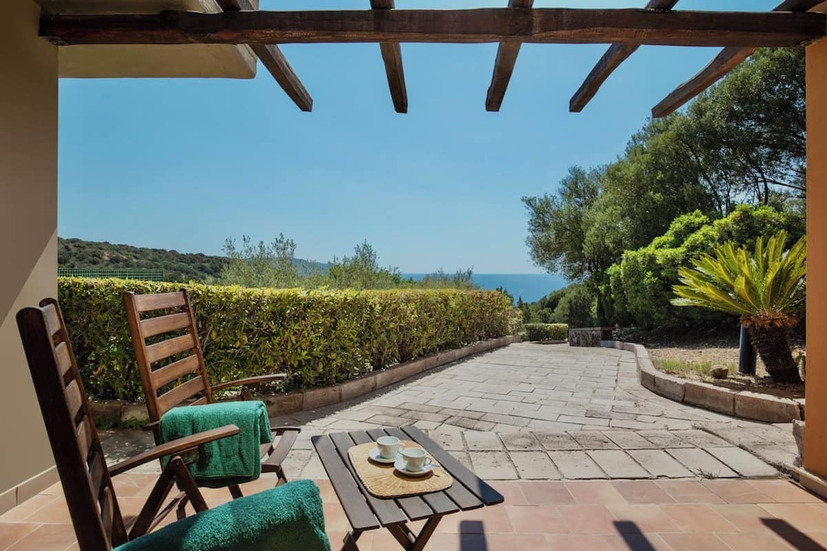 Une terrasse extérieure dotée d’une pergola en bois, une table avec deux tasses et leurs soucoupes, et une chaise sur laquelle est posée une serviette verte.