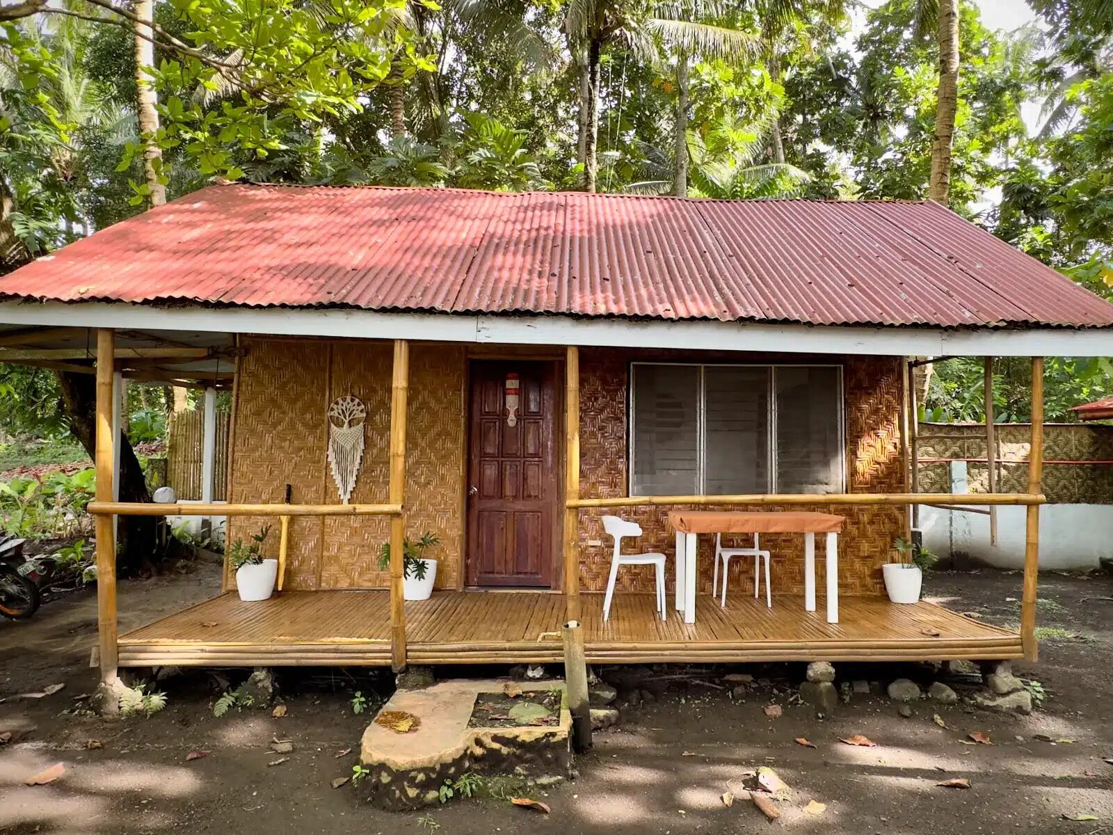 A rustic wooden house with a corrugated metal roof, a wooden porch with two white chairs, and a large window.