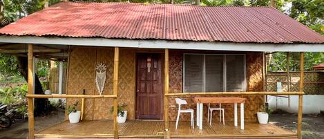 A rustic wooden house with a corrugated metal roof, a wooden porch with two white chairs, and a large window.