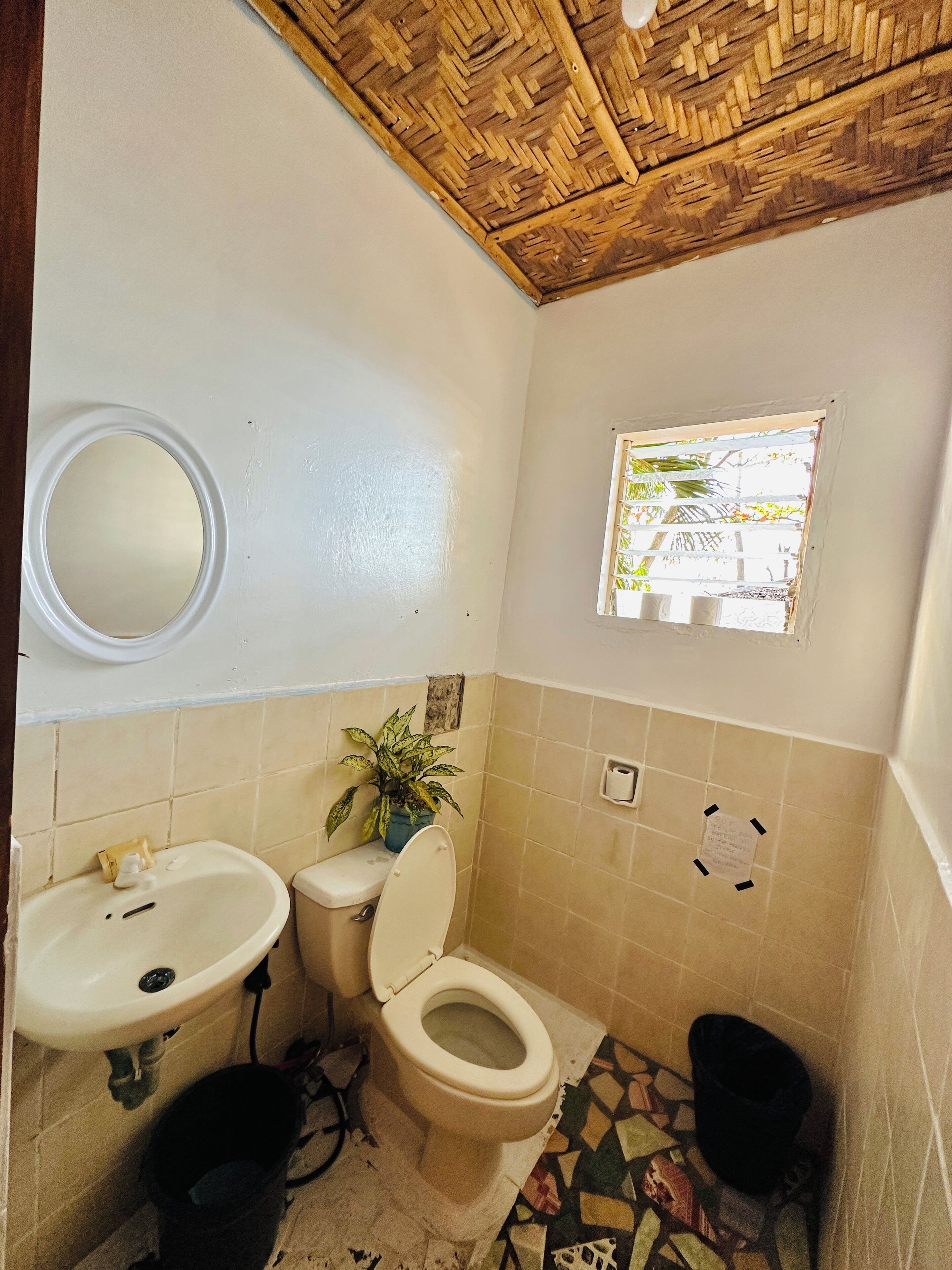 A bathroom with a wooden ceiling, a toilet, a sink, and a window with a view of greenery.