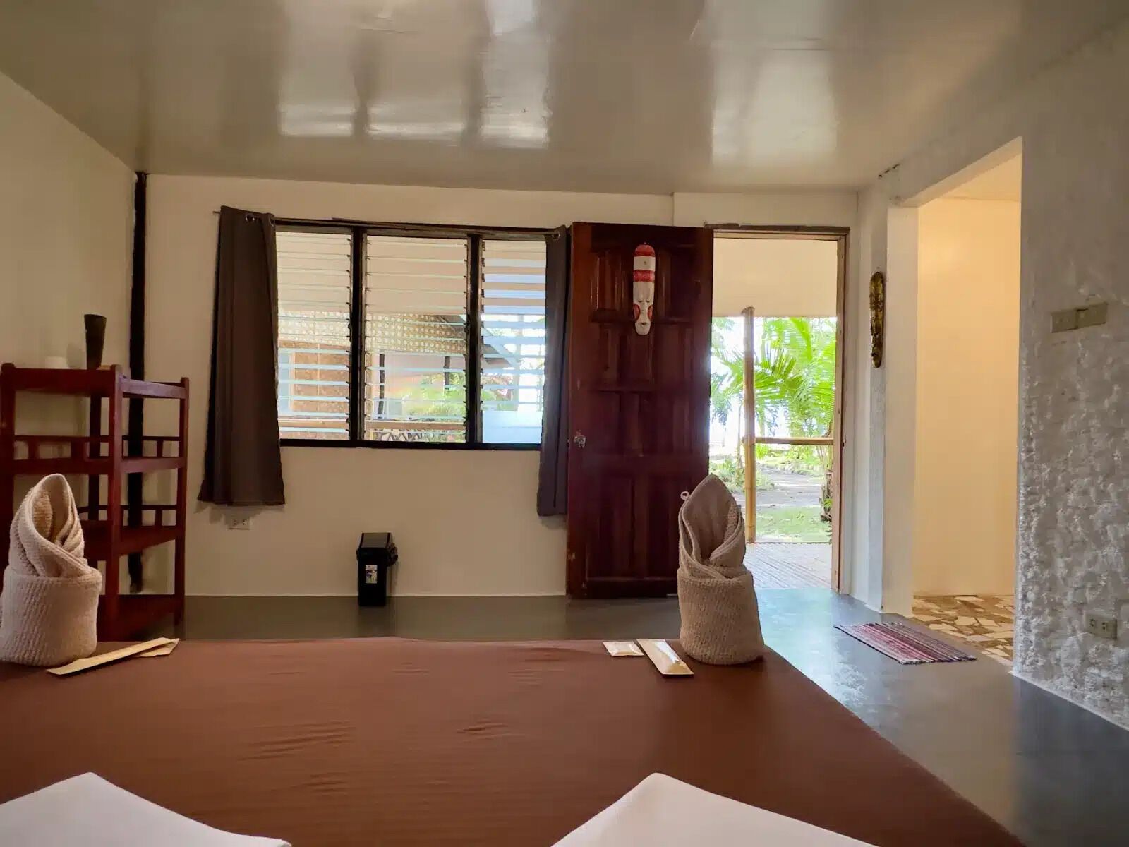 Una habitación con una cama de madera, una mesita pequeña, una silla, una ventana con persianas y una puerta que da al exterior.