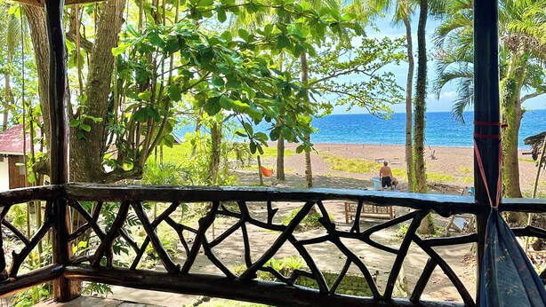 Una terraza de madera con techo tejido, con vistas a una playa con palmeras y una vista despejada del océano.