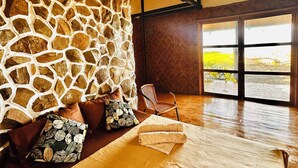Una habitación con pared de piedra, vigas de madera, un ventanal grande y una cama con almohadas y una colcha.