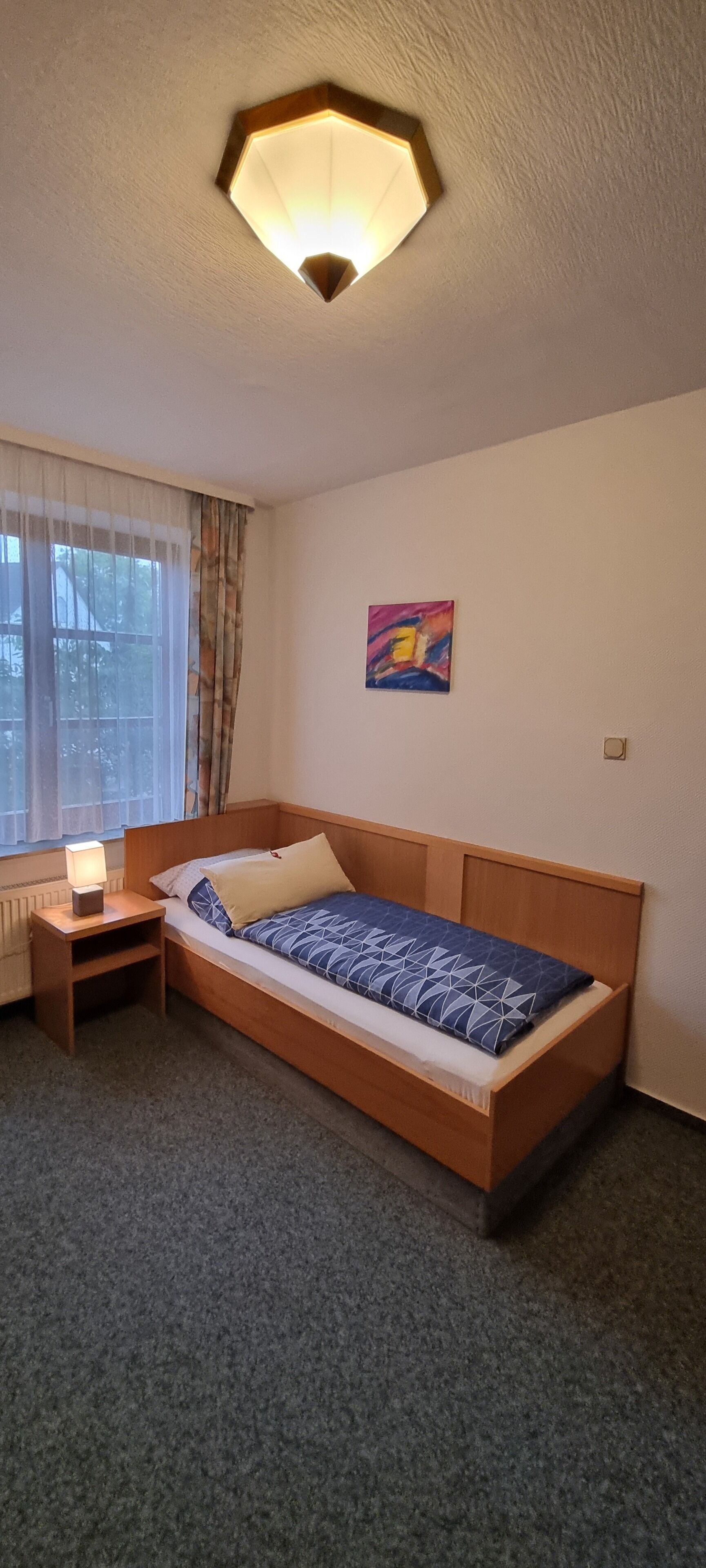 Una cama individual con cabecera de madera, una mesita de noche con lámpara, una ventana con cortinas translúcidas y un cuadro abstracto de colores en la pared.