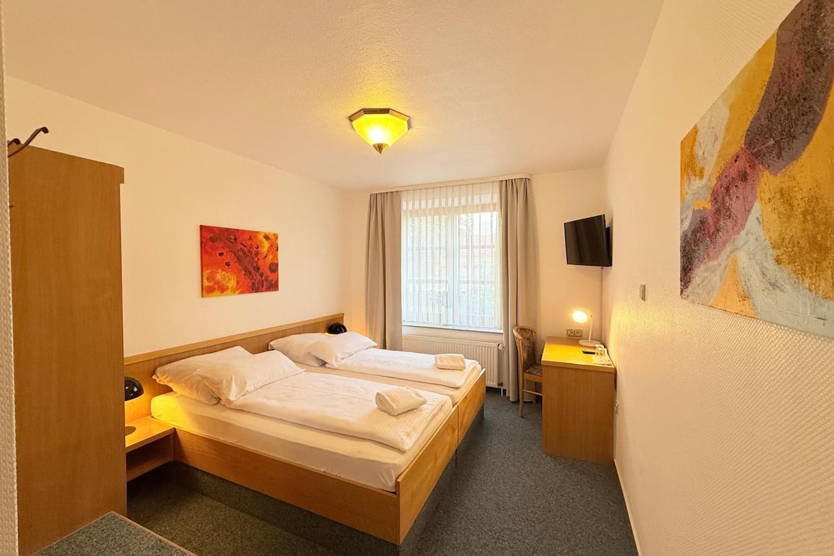 A hotel room with a double bed, bedside tables, a desk with a chair, a television, and a window with curtains.