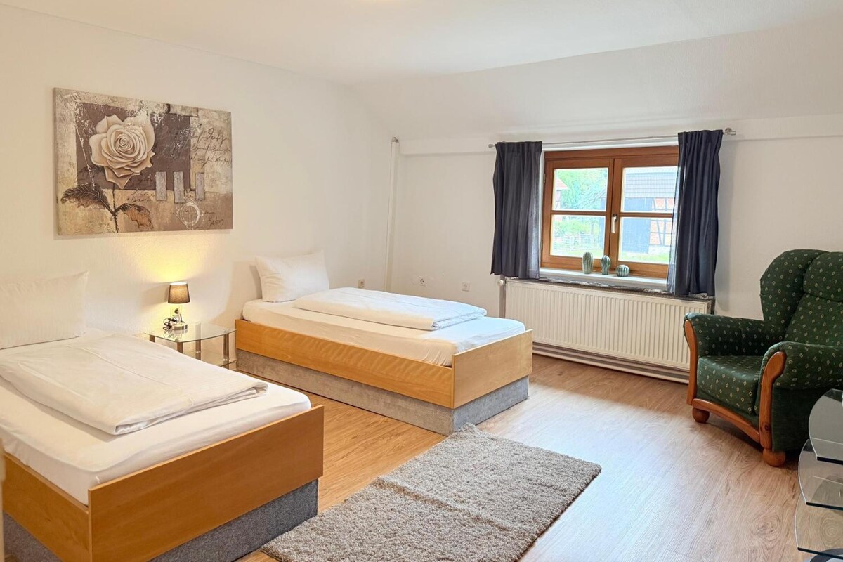 A hotel room with two beds, a window with curtains, a radiator, a green armchair, and a painting on the wall.