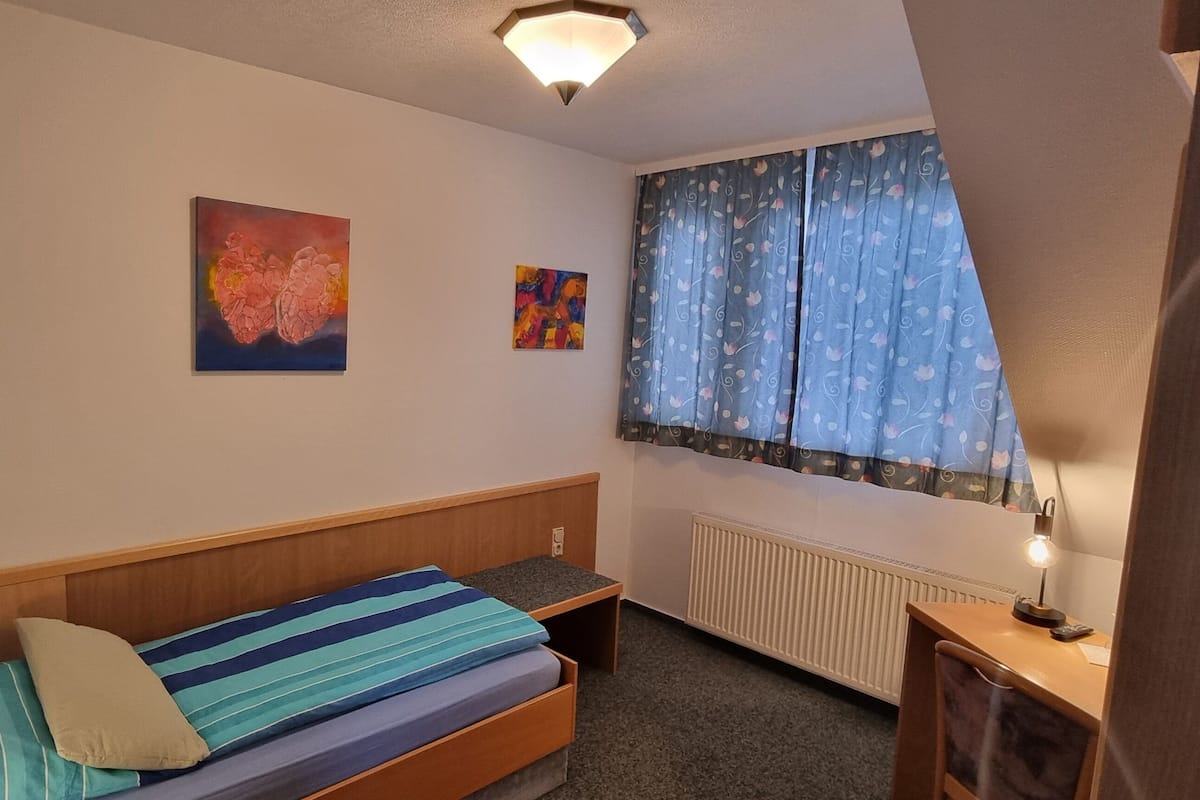A small bedroom with a single bed, a nightstand, a desk with a lamp, and a window with curtains.
