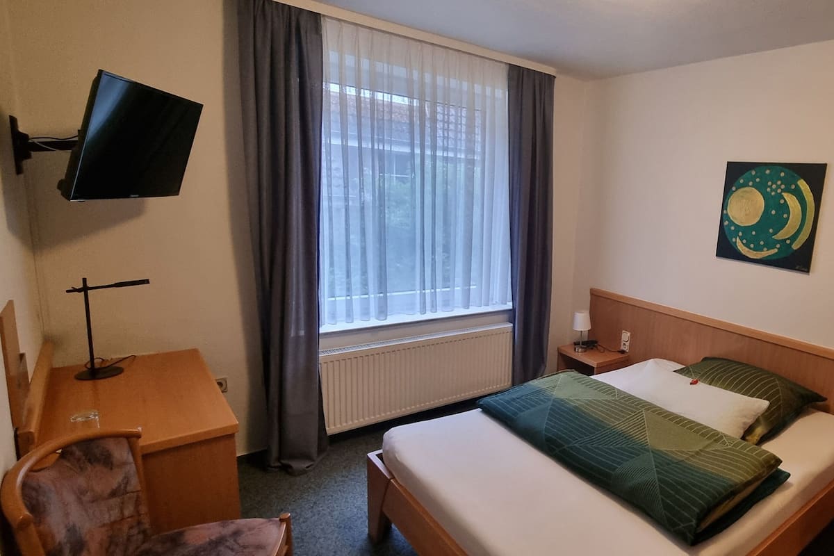 A hotel room with a bed, a desk, a chair, a TV mounted on the wall, a window with curtains, and a painting on the wall.