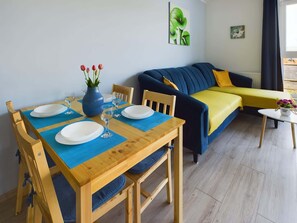 A dining area with a wooden table set for two, a blue vase with flowers, and a blue mat. A dark blue sofa with yellow cushions is against the wall.