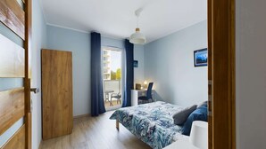Une chambre à coucher avec un lit, un bureau et une chaise. Il y a une fenêtre avec des rideaux, une porte et un ventilateur de plafond.