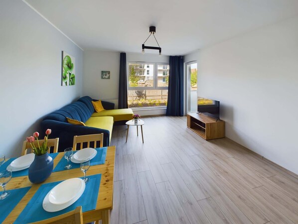 A modern living room with a sofa, a dining table set for two, a TV, and a large window with curtains.