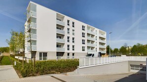 Un immeuble résidentiel moderne à plusieurs étages, doté de balcons, d’un espace de stationnement couvert et d’un jardin aménagé.