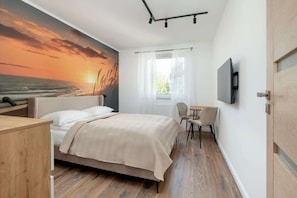 Un dormitorio con cama, escritorio, silla, televisor y un mural con un atardecer.