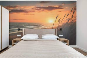 A bedroom with a large bed, two bedside tables, and a wall mural of a sunset over the ocean.