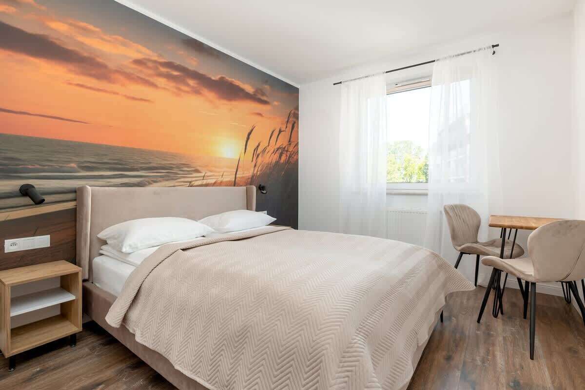 A bedroom with a bed, a desk, and a chair, a window with curtains, and a wall mural of a sunset.