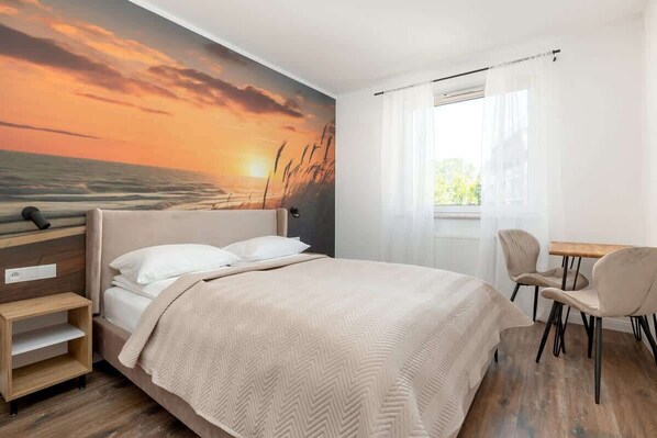 A bedroom with a bed, a desk, and a chair, a window with curtains, and a wall mural of a sunset.