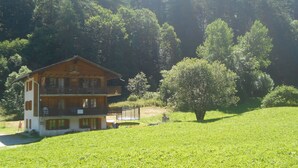 Una casa de madera de dos pisos con un balcón, rodeada por un campo cubierto de hierba y un bosque denso.