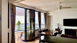 A dining area with a table and chairs, a TV mounted on the wall, and a balcony with patterned curtains.