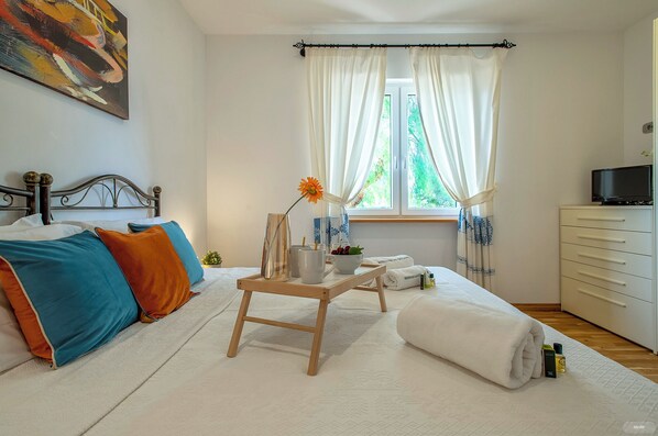 A bedroom with a bed, bedside table, curtains, a window, a TV, and a dresser.
