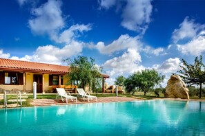A swimming pool with lounge chairs, a stone building with a tiled roof, and a clear blue sky.